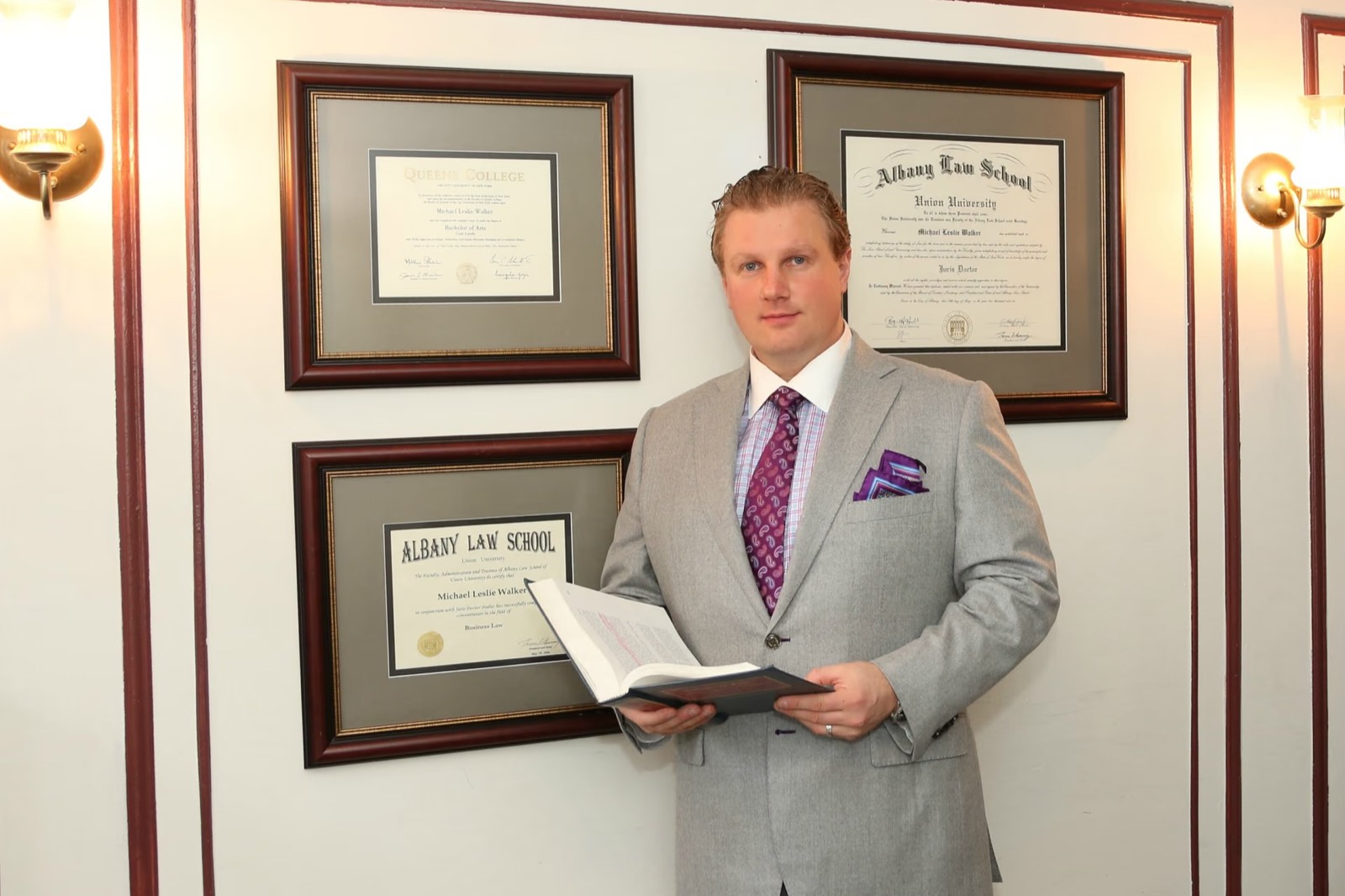 Attorney Michael L. Walker holding a legal treatise beside his Queens College and Albany Law School diplomas
